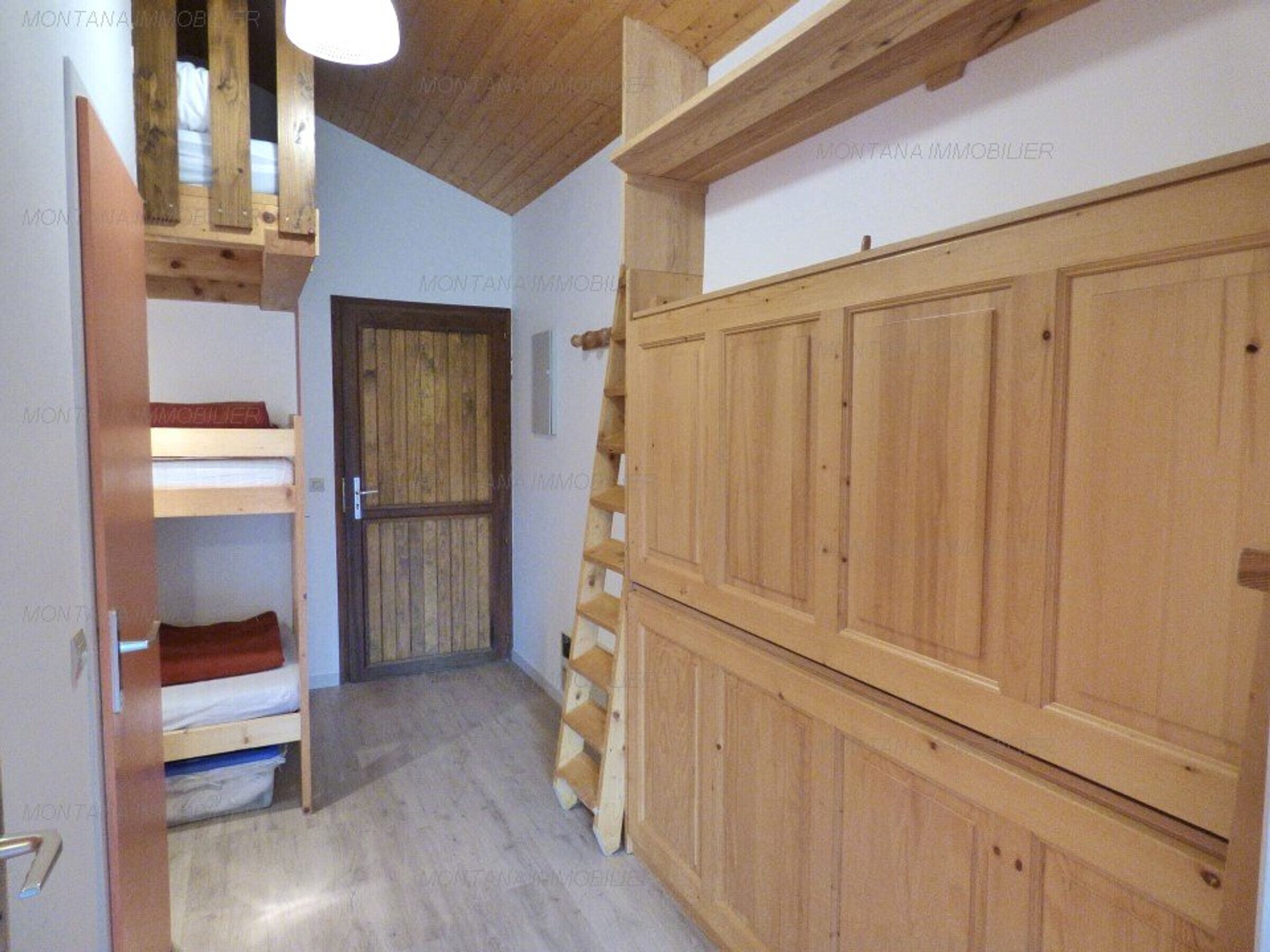 A narrow hallway with wooden walls and a door. There is a wooden ladder leaning against the wall and a bunk bed with a red blanket.