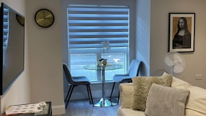 A modern living room with a glass table, two chairs, a sofa, a clock, a framed artwork, and a window with blinds. - Catharine House by condokeeper (Cambridge)
