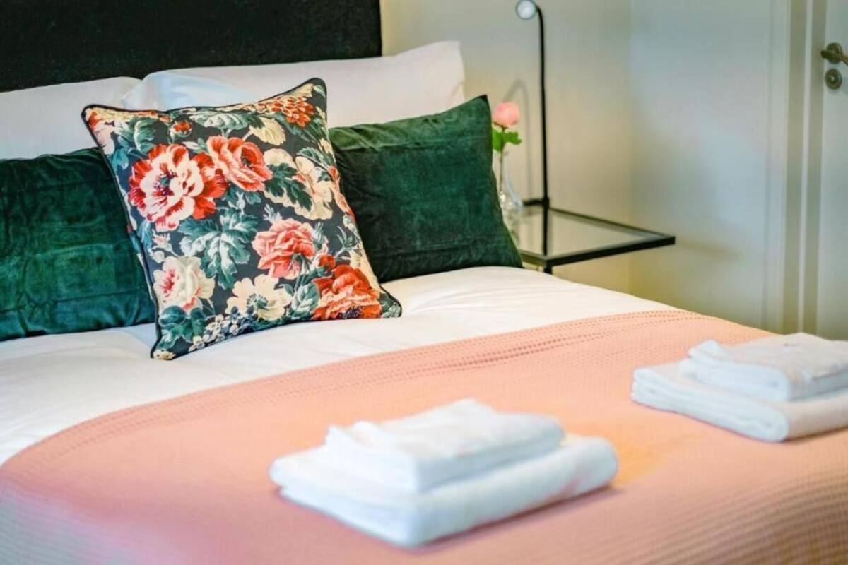 A neatly made bed with floral and solid green pillows, a pink bedspread, and folded white towels on a bedside table.