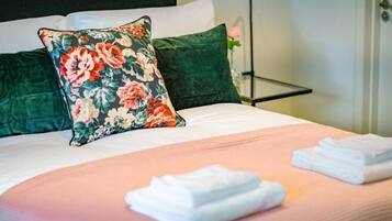 A neatly made bed with floral and solid green pillows, a pink bedspread, and folded white towels on a bedside table.