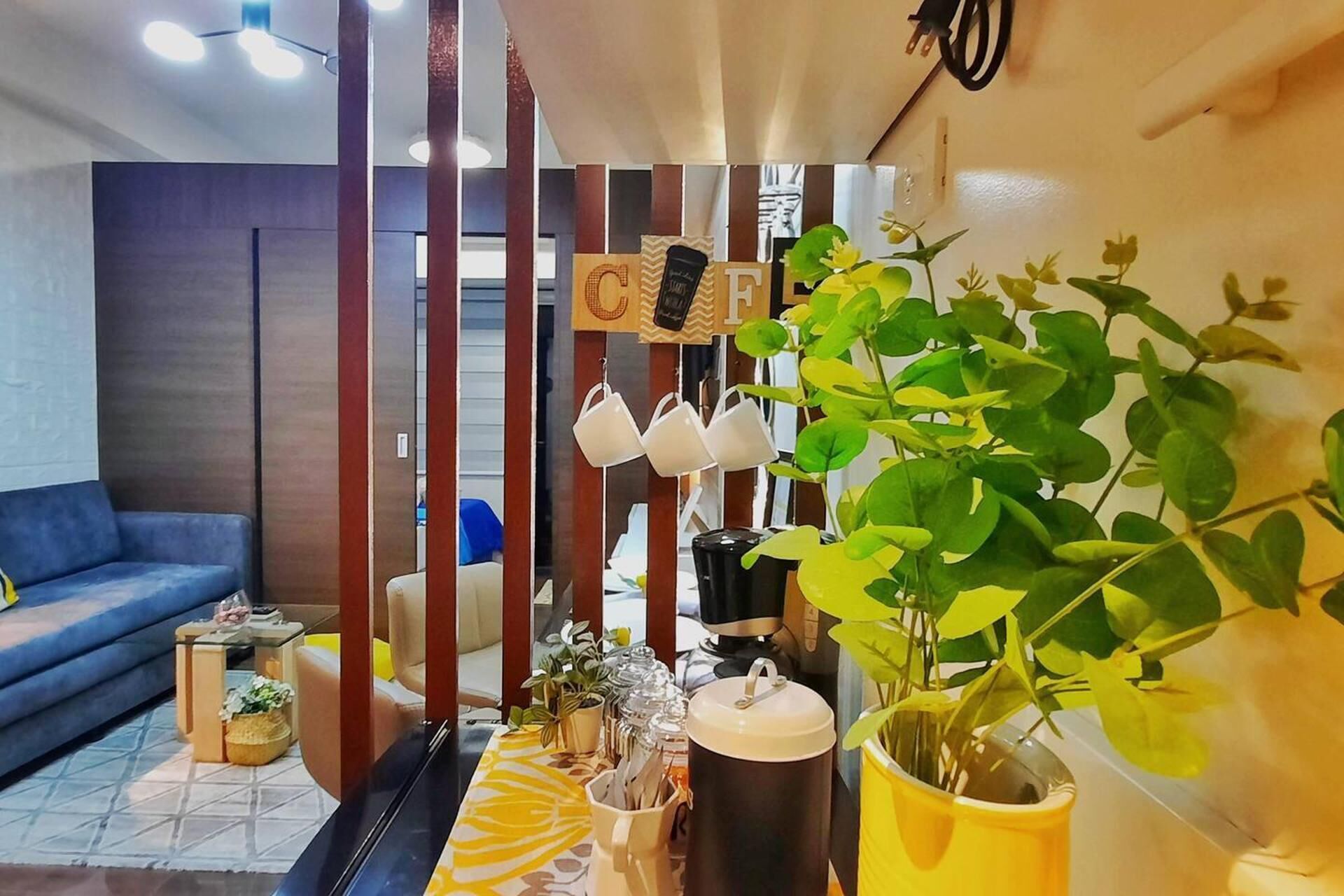 A living room with a blue sofa, a coffee table with a yellow patterned tablecloth, a potted plant, and a decorative wall with letters and a clock.
