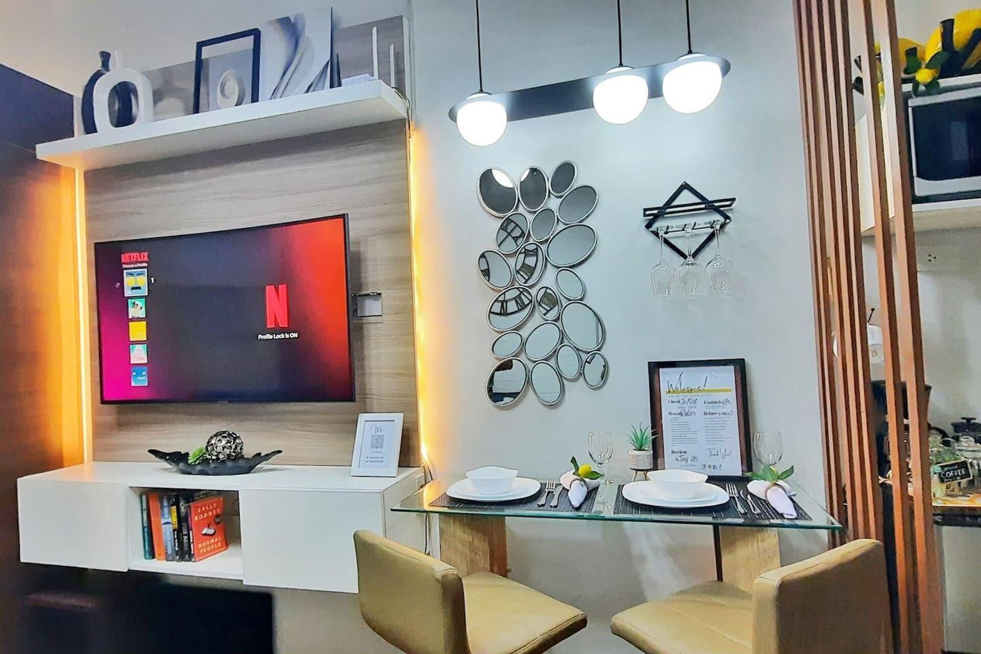 A modern hotel lobby with a flat-screen TV displaying a Netflix interface, a glass-top dining table set for two, and a wall-mounted mirror with a geometric design.