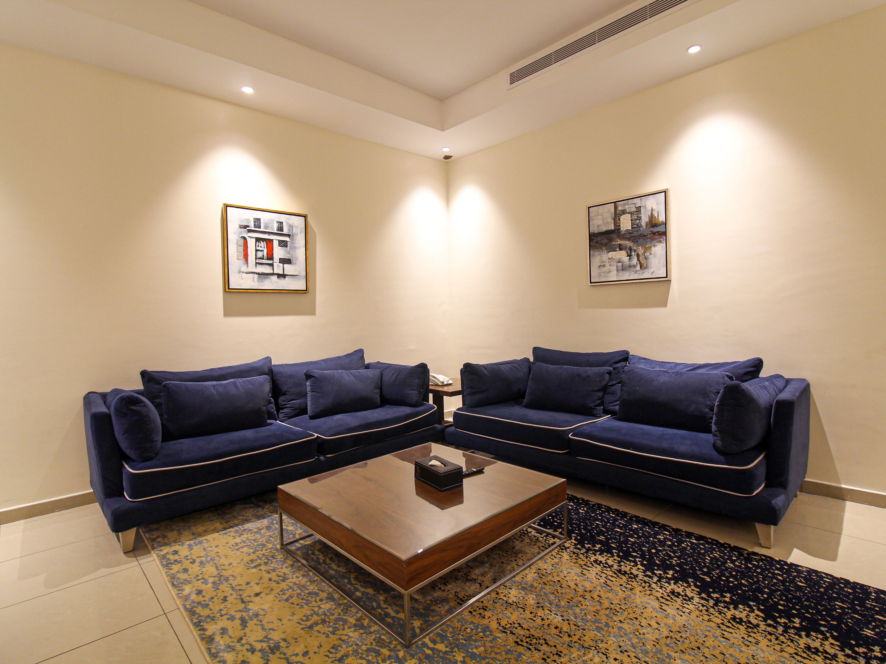A room with two dark blue sofas arranged in an L-shape around a wooden coffee table. There are two framed artworks on the walls.