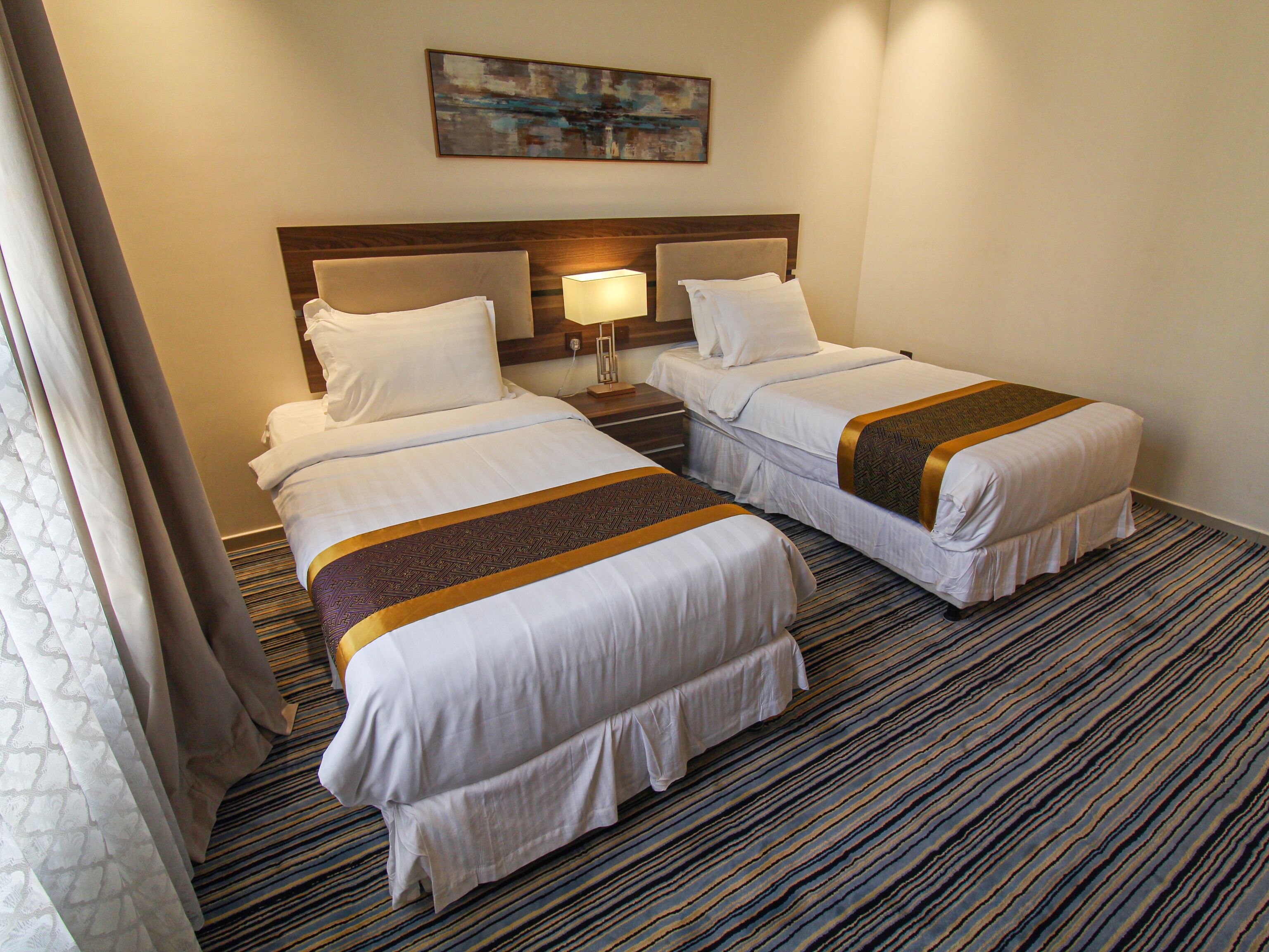 A hotel room with two beds, a headboard, a lamp, and a painting on the wall.