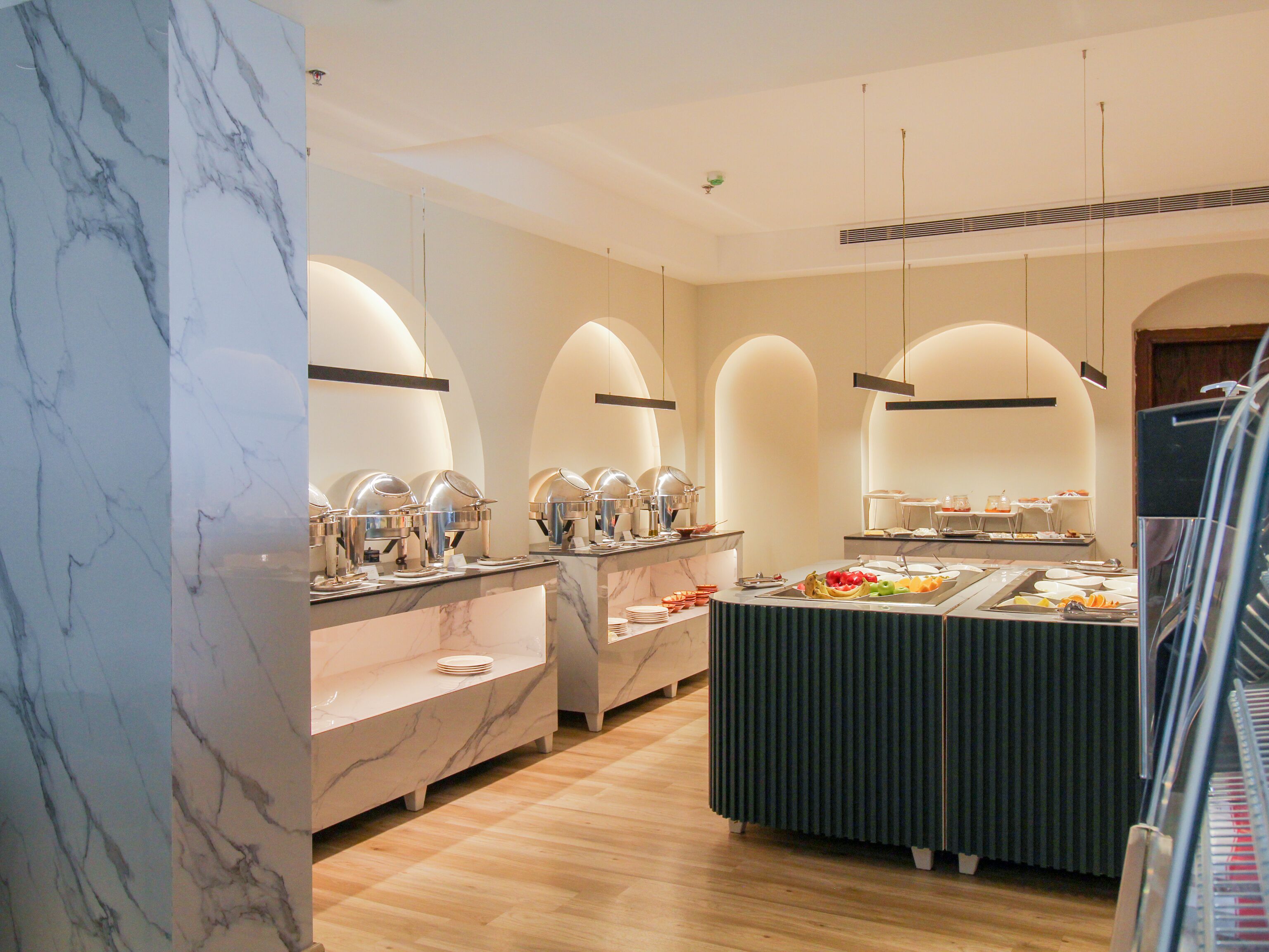 Uma área de buffet com bancadas de mármore, diversas estações de serviço e uma mesa central com frutas.