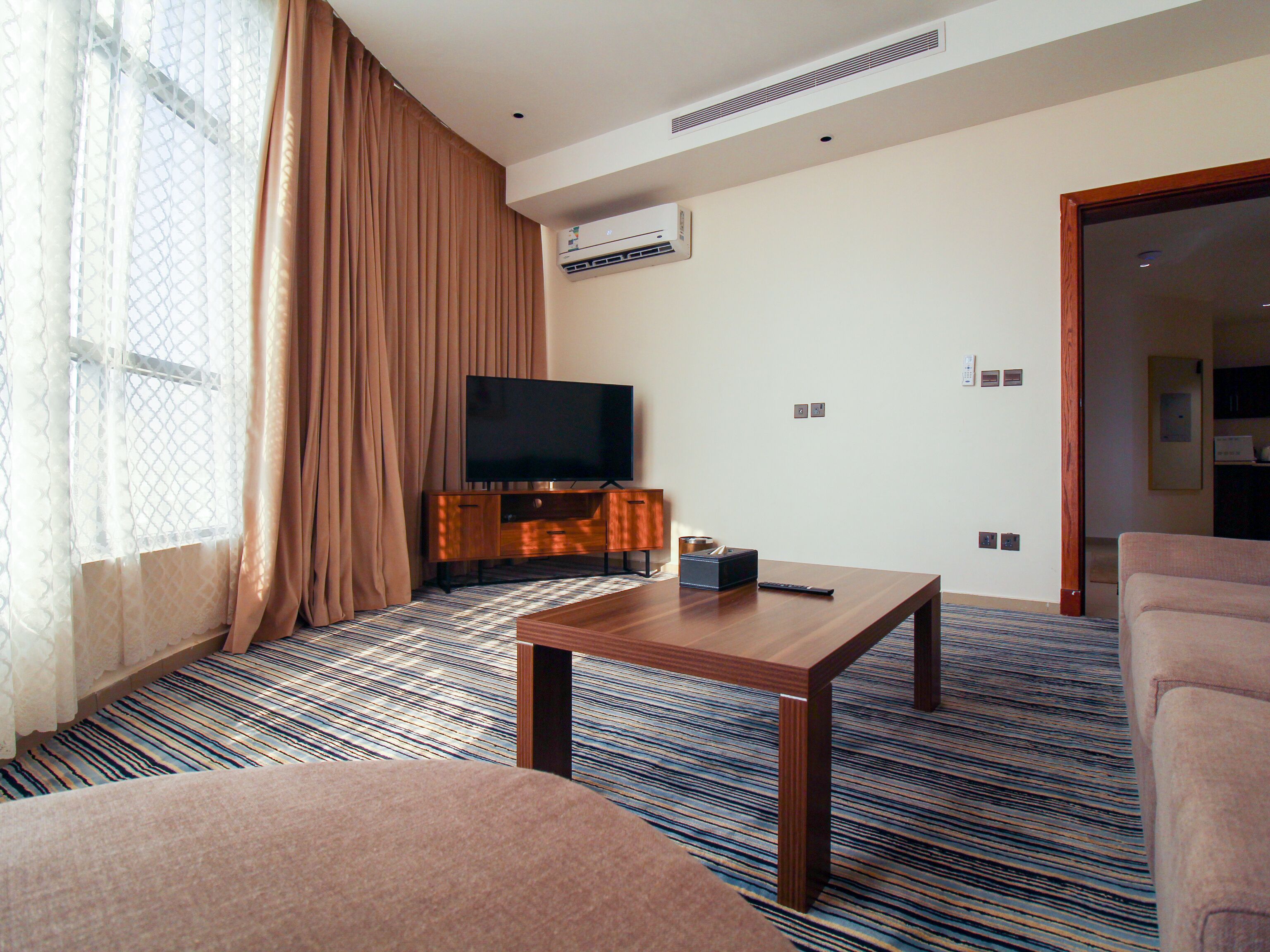 A hotel room with a flat-screen TV on a wooden cabinet, a coffee table, a sofa, and a patterned carpet.