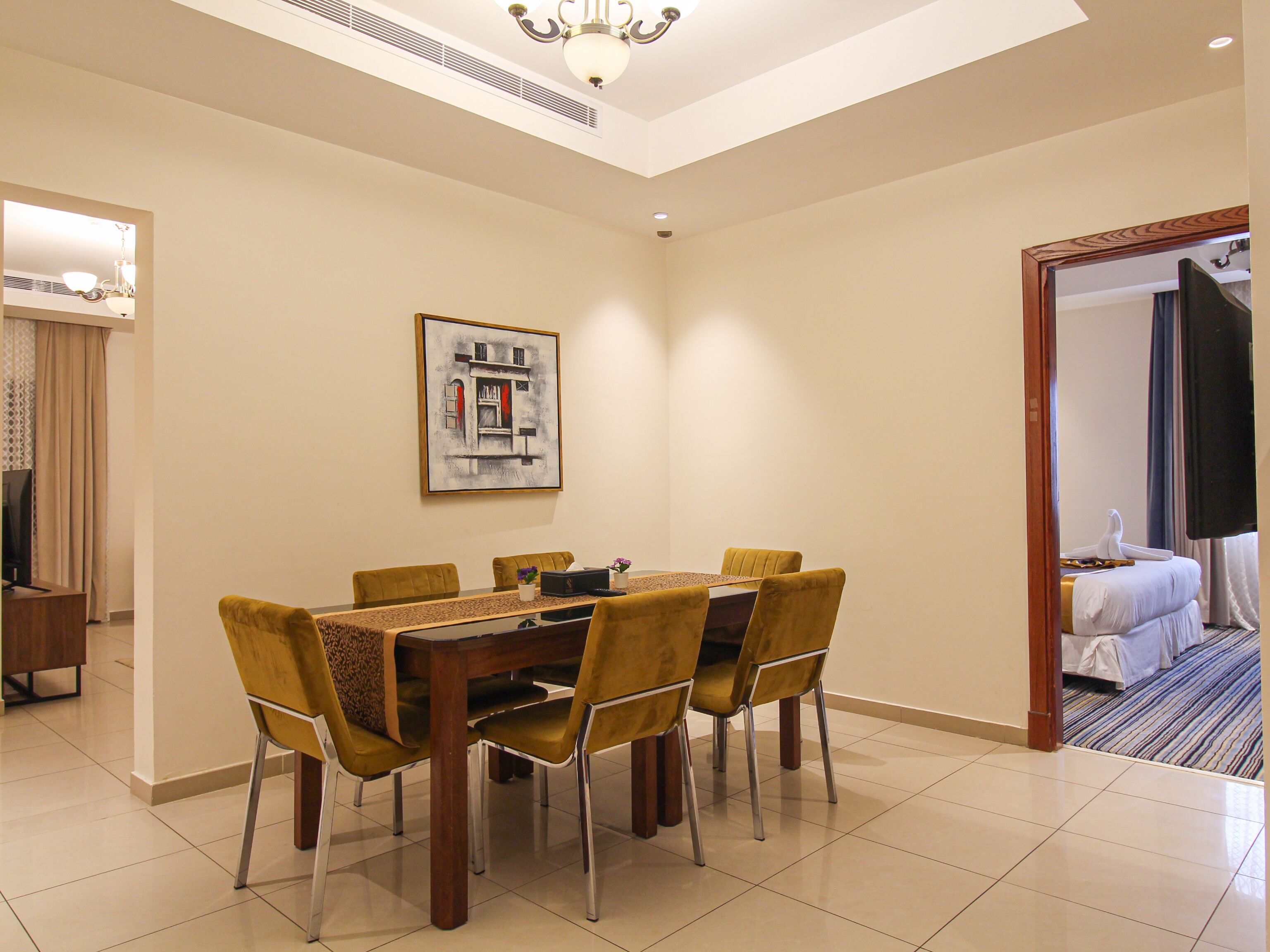 A hotel dining area with a round table, six chairs, and a painting on the wall.