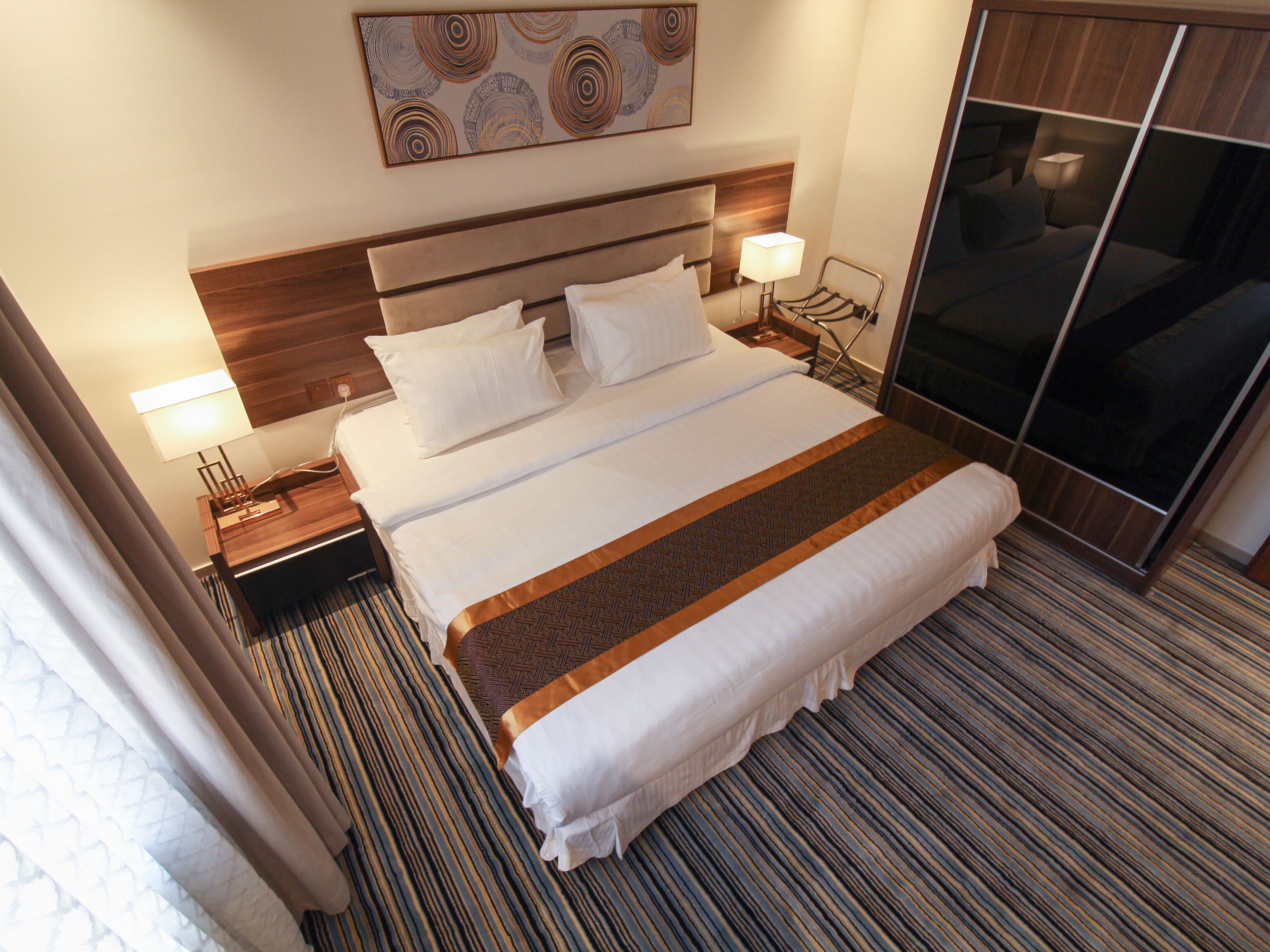 Habitación de hotel con una cama grande, dos mesitas de noche con lámparas, un cabecero, una obra de arte en la pared y un armario corredero.