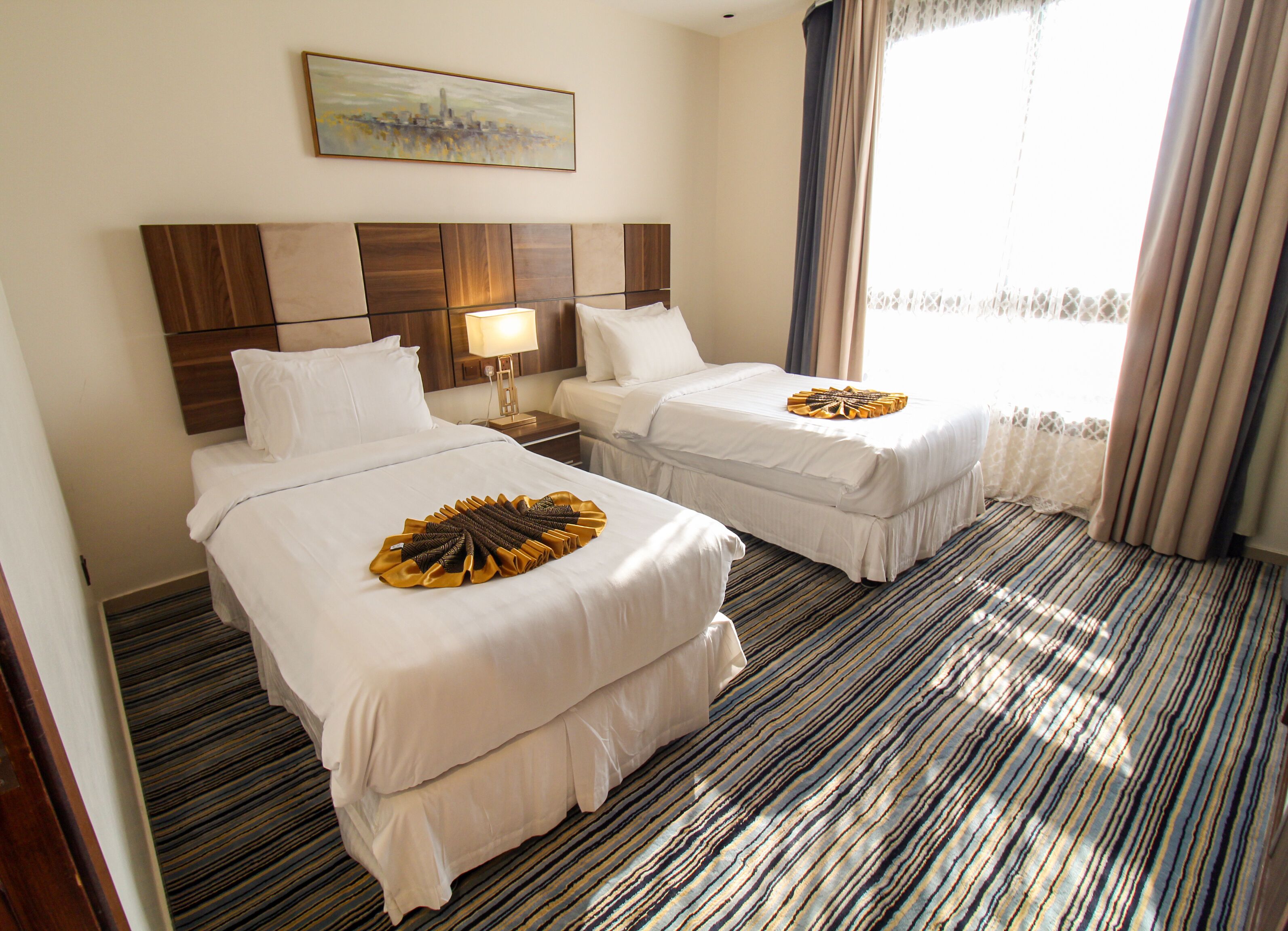 Ein Hotelzimmer mit zwei Betten, einem Kopfteil mit Stadtbild-Gemälde, einem Nachttisch mit Lampe und einem Streifenmuster-Teppich.