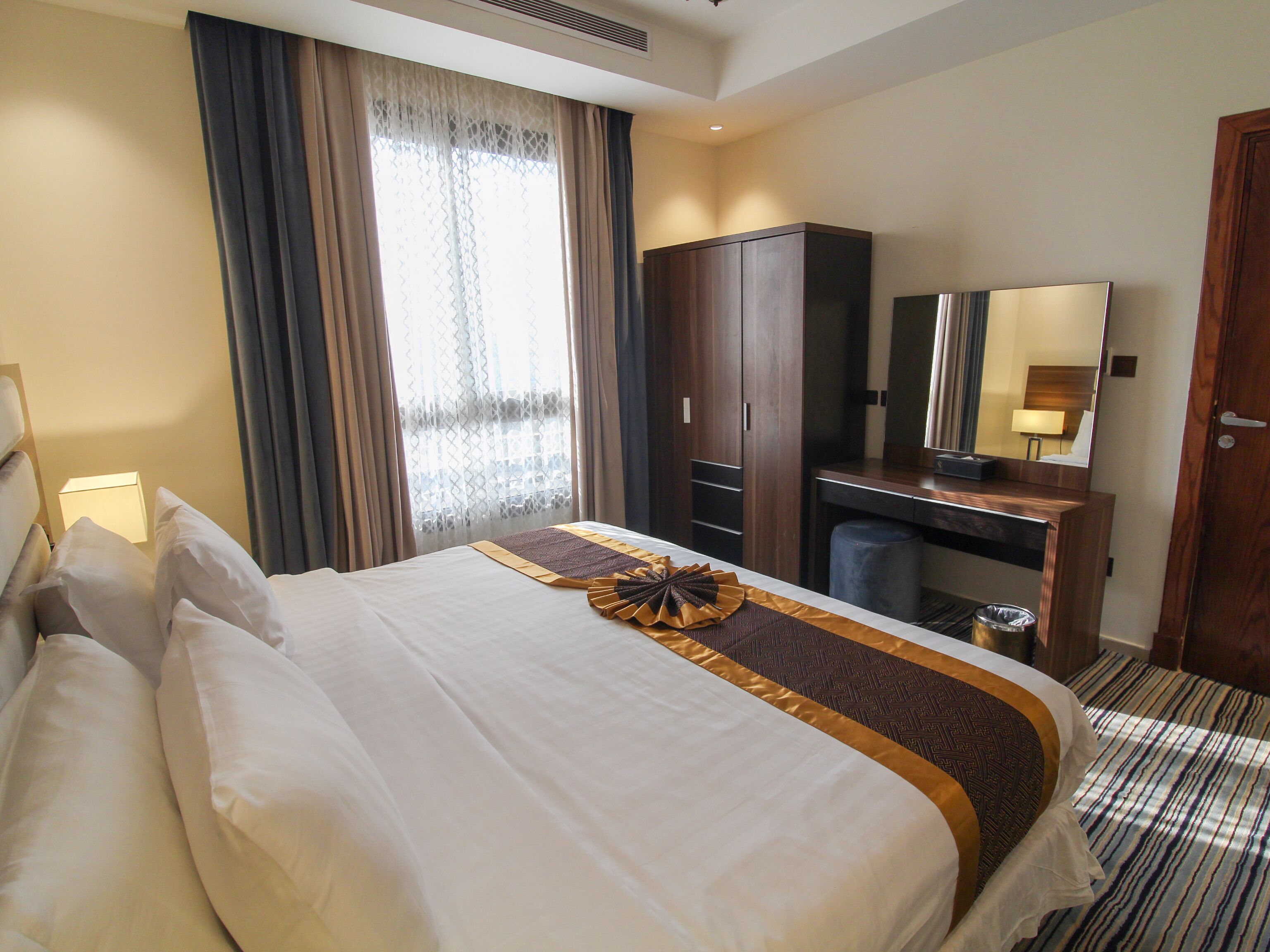 A hotel room with a neatly made bed, a desk with a mirror, and a wardrobe.