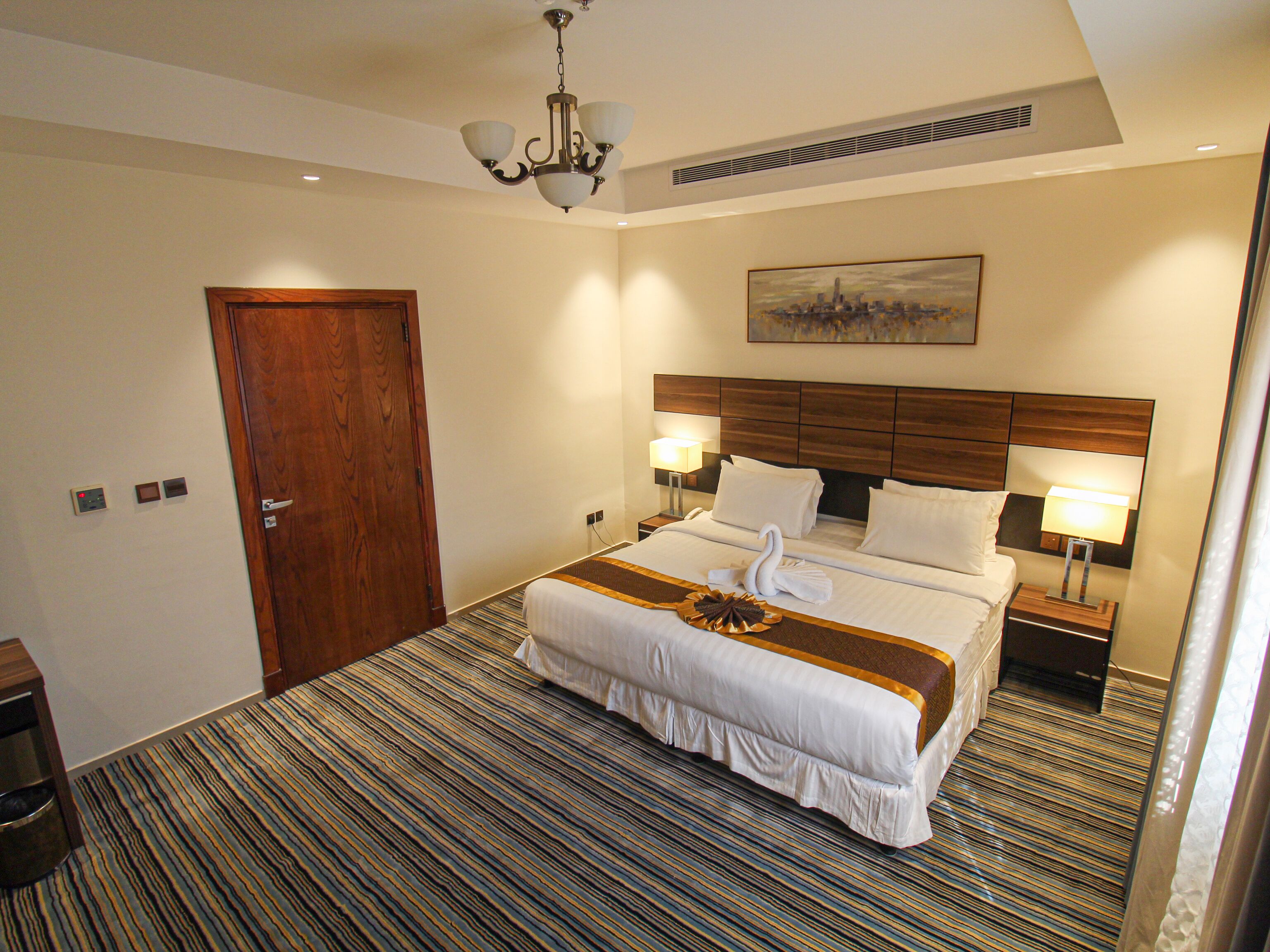 Een hotelkamer met een groot bed, twee bedlampen, een houten hoofdeinde, een schilderij aan de muur en een kamerbreed tapijt met dessin.