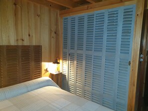A bedroom with wooden walls, a bed with a white quilt, a nightstand with a lamp, and a blue louvered closet. - Cabane Ferret Capienne, a 5-minute walk from the lake or the ocean (Lège-Cap-Ferret)