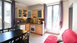 Cozinha compacta com armários de madeira, micro-ondas e máquina de café. Há uma mesa de jantar com cadeiras e um sofá vermelho na sala de estar.