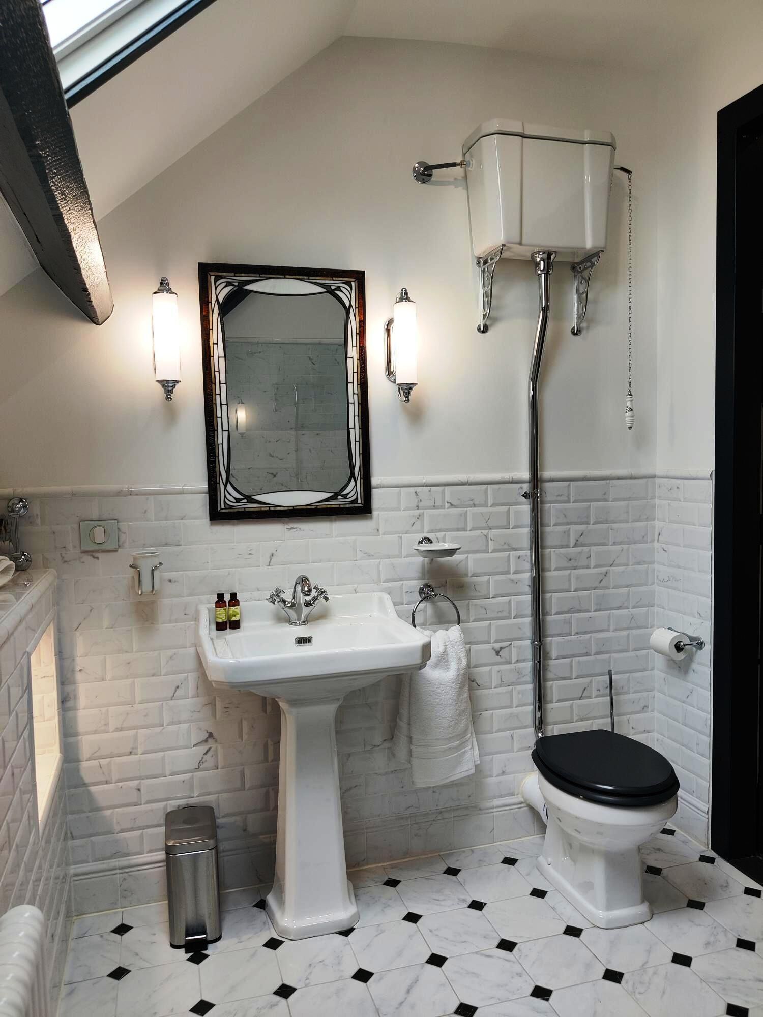 A bathroom with a white pedestal sink, a black toilet, a white toilet paper holder, a white trash can, and a mirror with a black frame.