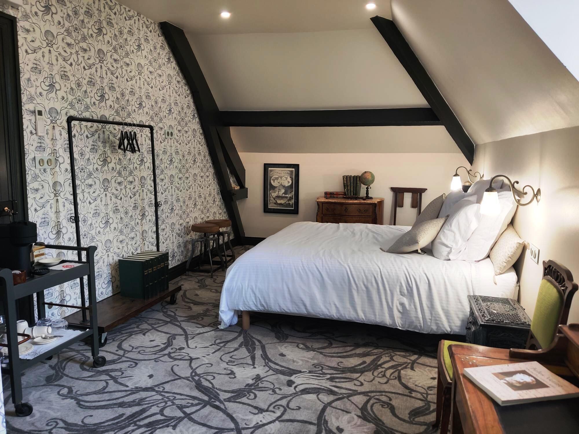 A bedroom with a high ceiling, a bed with white linens, a wooden nightstand, a small table with a lamp, and a patterned carpet.