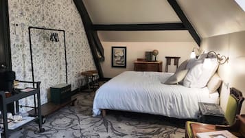 A bedroom with a high ceiling, a bed with white linens, a wooden nightstand, a small table with a lamp, and a patterned carpet.