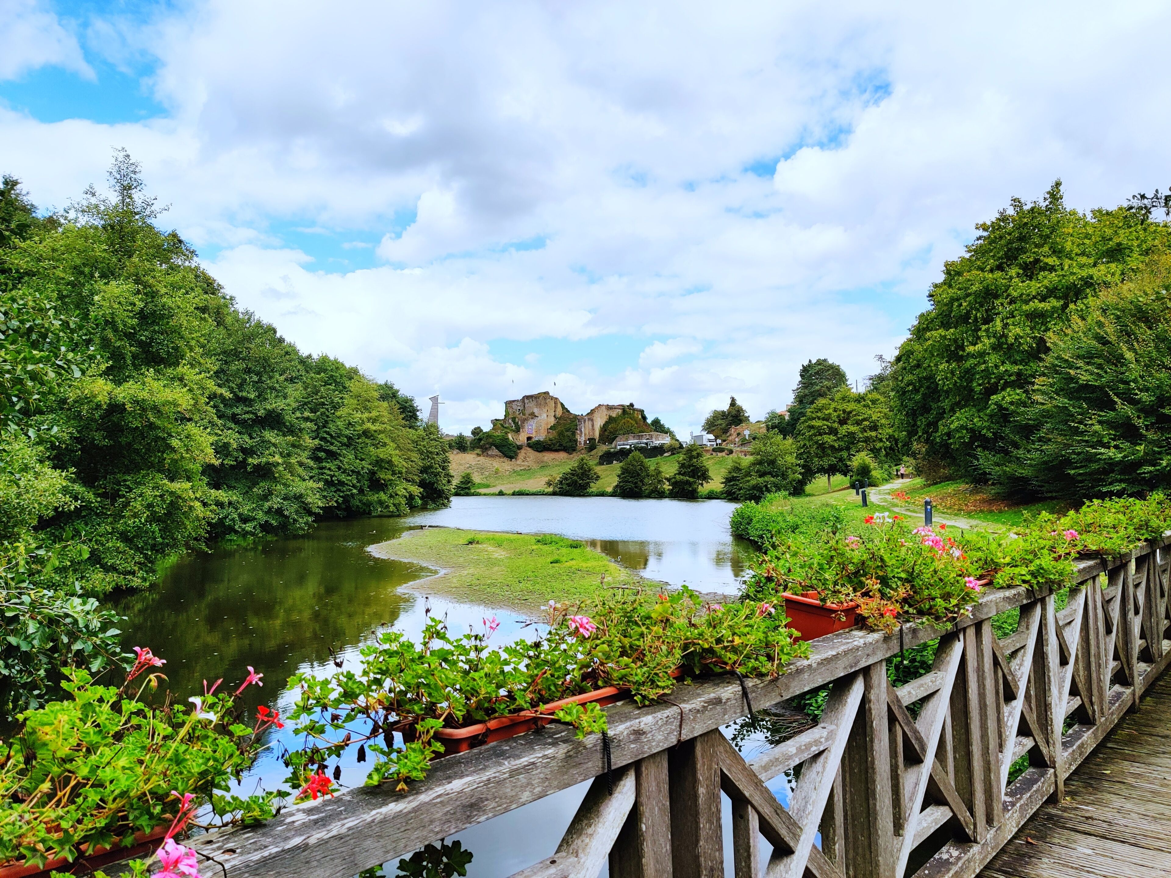 Un puente de madera con macetas y flores, que conduce a un río tranquilo con exuberante vegetación y un castillo al fondo.