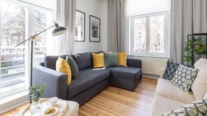 A modern living room with a sectional sofa, a floor lamp, and a view of the city through large windows. - London - Cozy City Stay by Lion Apartments Sopot (Sopot)