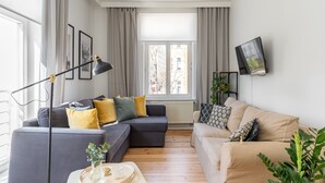 A modern living room with a grey sectional sofa, a beige sofa, and a coffee table with a vase. There is a floor lamp, a TV mounted on the wall, and a window with curtains. - London - Cozy City Stay by Lion Apartments Sopot (Sopot)