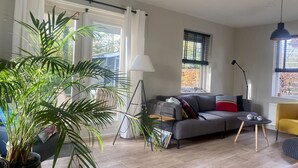 Una sala de estar con un sofá gris, un sillón amarillo, una lámpara de pie y una planta grande en maceta.