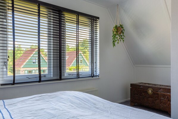 A bedroom with a bed, a chest of drawers, a window with blinds, and a hanging plant.