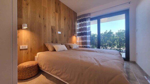 Un dormitorio moderno con una cama grande, paneles de madera en la pared, un ventanal con vistas y un taburete redondo de mimbre.