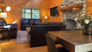 A cozy log cabin interior with a stone fireplace, leather sofas, and a dining area. - LUXE TWO | ANTLER'S GROVE *HOCKING HILLS* CLOSE TO EVERYTHING! HOT TUB, FIREPLACE,GAME ROOM,FIRE PIT (Laurelville)