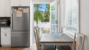 Cocina compacta con refrigerador, microondas y armarios de madera. Un comedor con sillas se encuentra cerca de un amplio ventanal con persianas.