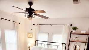 Een slaapkamer met een bed, een bankje, een plafondventilator en een plank met decoratieve spullen.