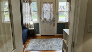 A hallway leading to a bedroom with a bed, a nightstand, a lamp, and a door with patterned curtains.