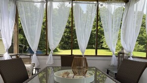A glass-top table with wicker chairs and a small table lamp, set against a backdrop of sheer white curtains and a view of a green landscape.