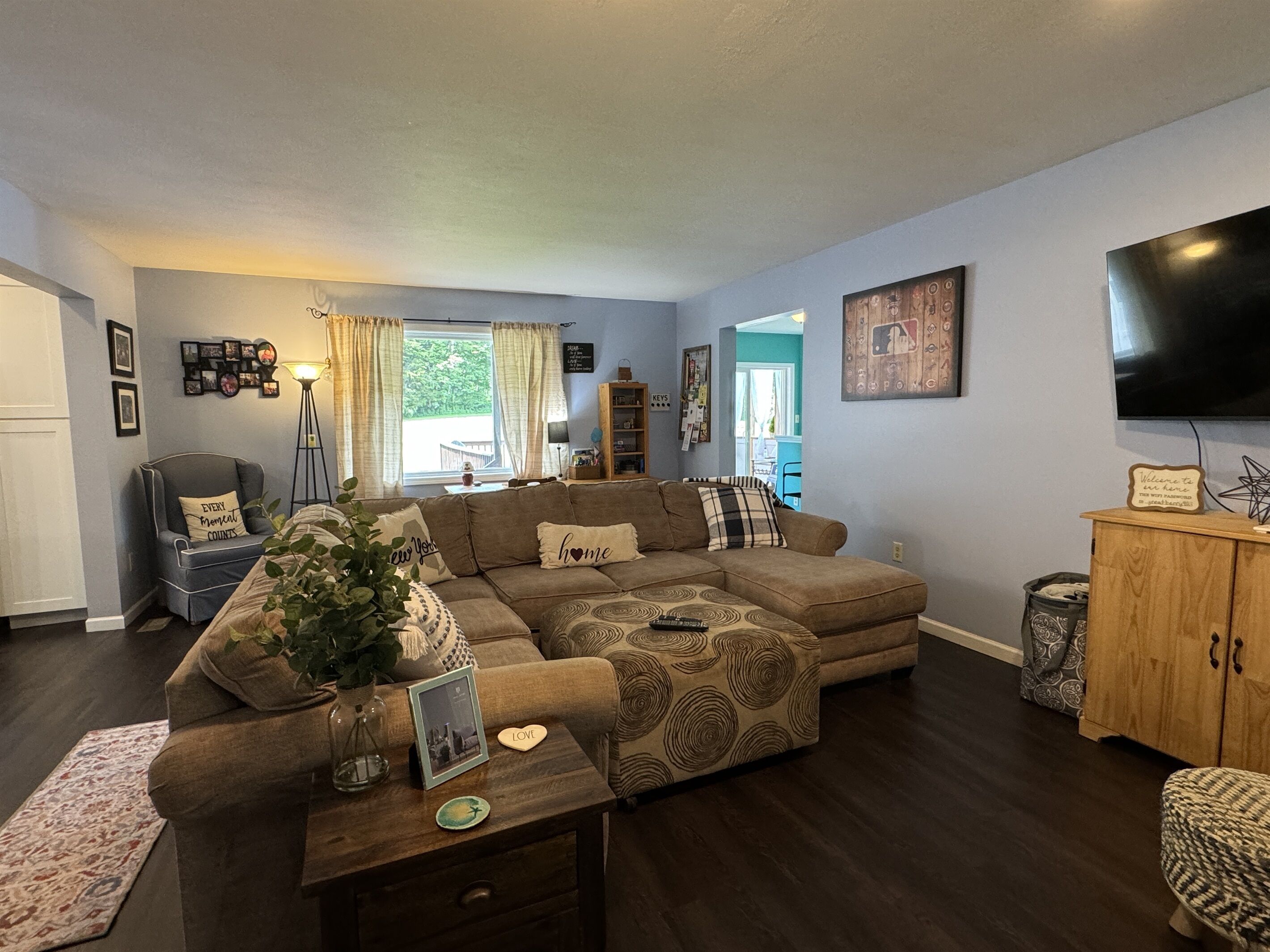 A living room with a sectional sofa, a coffee table, and a TV. There is a window with curtains, a bookshelf, and a cabinet.
