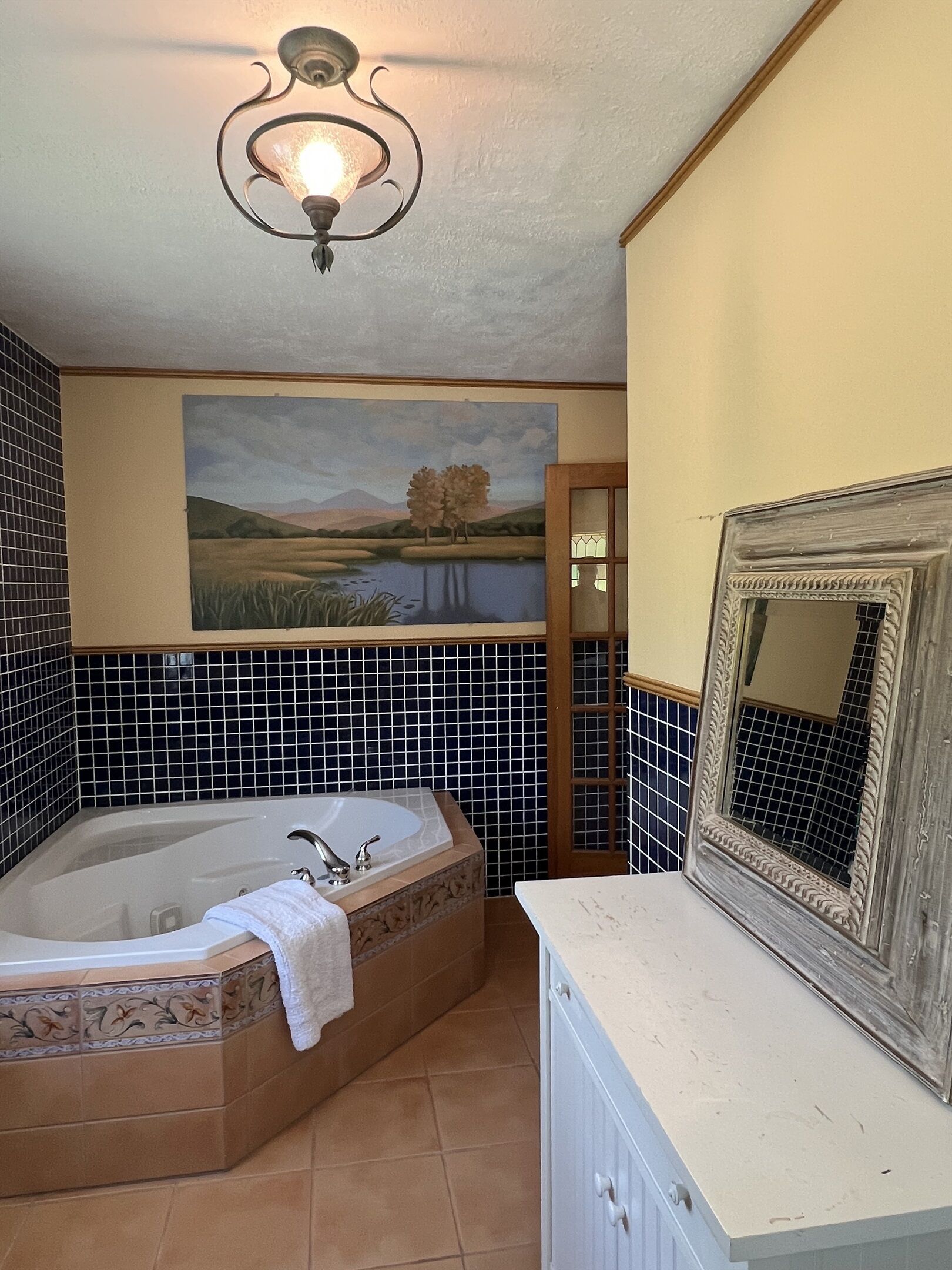 A bathroom with a large bathtub, a painting of a landscape, and a mirror with a decorative frame.