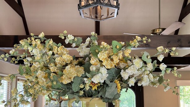Zona pranzo con un grande vaso di fiori su un tavolo in vetro, travi in legno a soffitto e un lampadario.