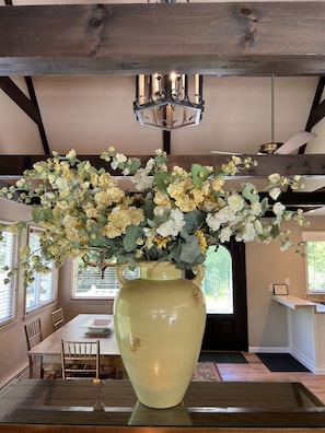Un coin repas agrémenté d’un grand vase rempli de fleurs posé sur une table en verre, de poutres en bois au plafond et d’un lustre.