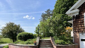 Un escalier en briques mène à une porte de garage blanche, le tout étant entouré d’une végétation luxuriante et d’arbres.