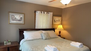 Cama bem arrumada com colcha florida, travesseiros e toalhas dobradas. Um criado-mudo com abajur e uma pequena planta. Duas fotos emolduradas na parede acima da cama.