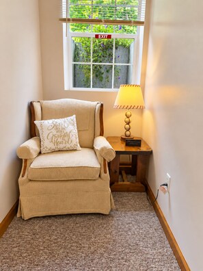 A corner room with a beige armchair, a small table with a lamp, and an exit sign above a window with greenery outside.
