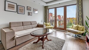 A modern living room with a sofa, a glass-top coffee table, and a view of buildings through large windows. - Lion Apartments - Parkur 2 (Sopot)
