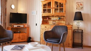 Una sala de estar acogedora con televisión, dos sillones, una mesa de centro con libros, un aparador de madera y una lámpara.