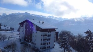 Un edificio de varios pisos con tejado cubierto de nieve, rodeado de árboles nevados y montañas al fondo.