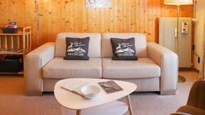 Una habitación acogedora con un muro de madera, un sofá blanco con dos cojines, una mesita con un cuenco y un libro, y un cartel en la pared.