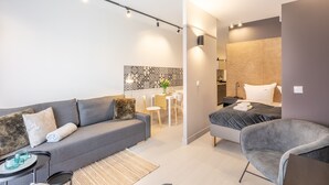 Un espace de vie moderne et bien éclairé, avec un canapé gris, un fauteuil gris et une petite table sur laquelle sont posés des verres. On y trouve un lit avec une couette grise et une table de chevet avec une lampe. La pièce présente une palette de couleurs neutres et un mur décoratif à motifs dans la partie cuisine.