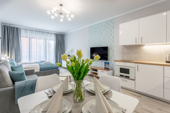 A modern living room with a sofa set, a dining table with set plates and napkins, a vase with flowers, and a kitchen area with cabinets and a TV.