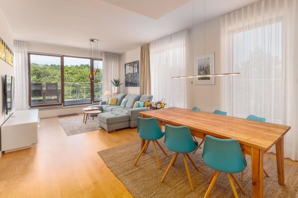 Uma sala de estar moderna com uma mesa de jantar de madeira, cadeiras verde-água, um sofá cinza e uma grande janela com vista para o verde.