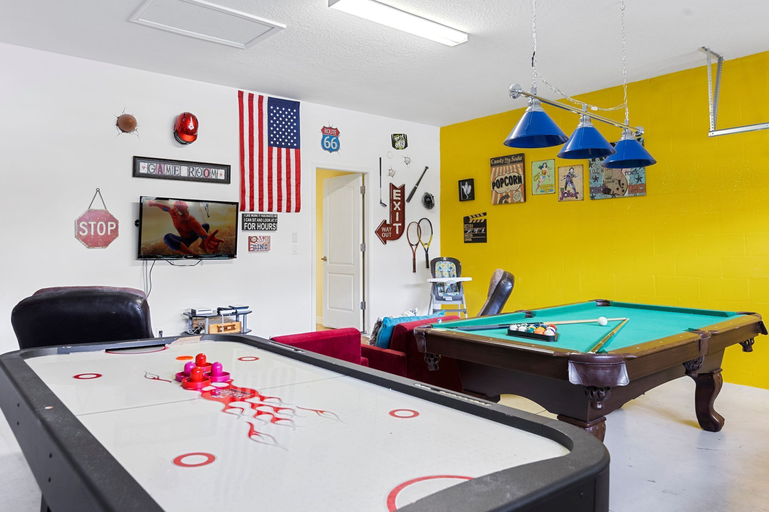 Une table de billard recouverte d’une nappe rouge et blanche, un queue de billard et une chaise rouge. Un téléviseur est fixé au mur, et une porte donne accès à une autre pièce.