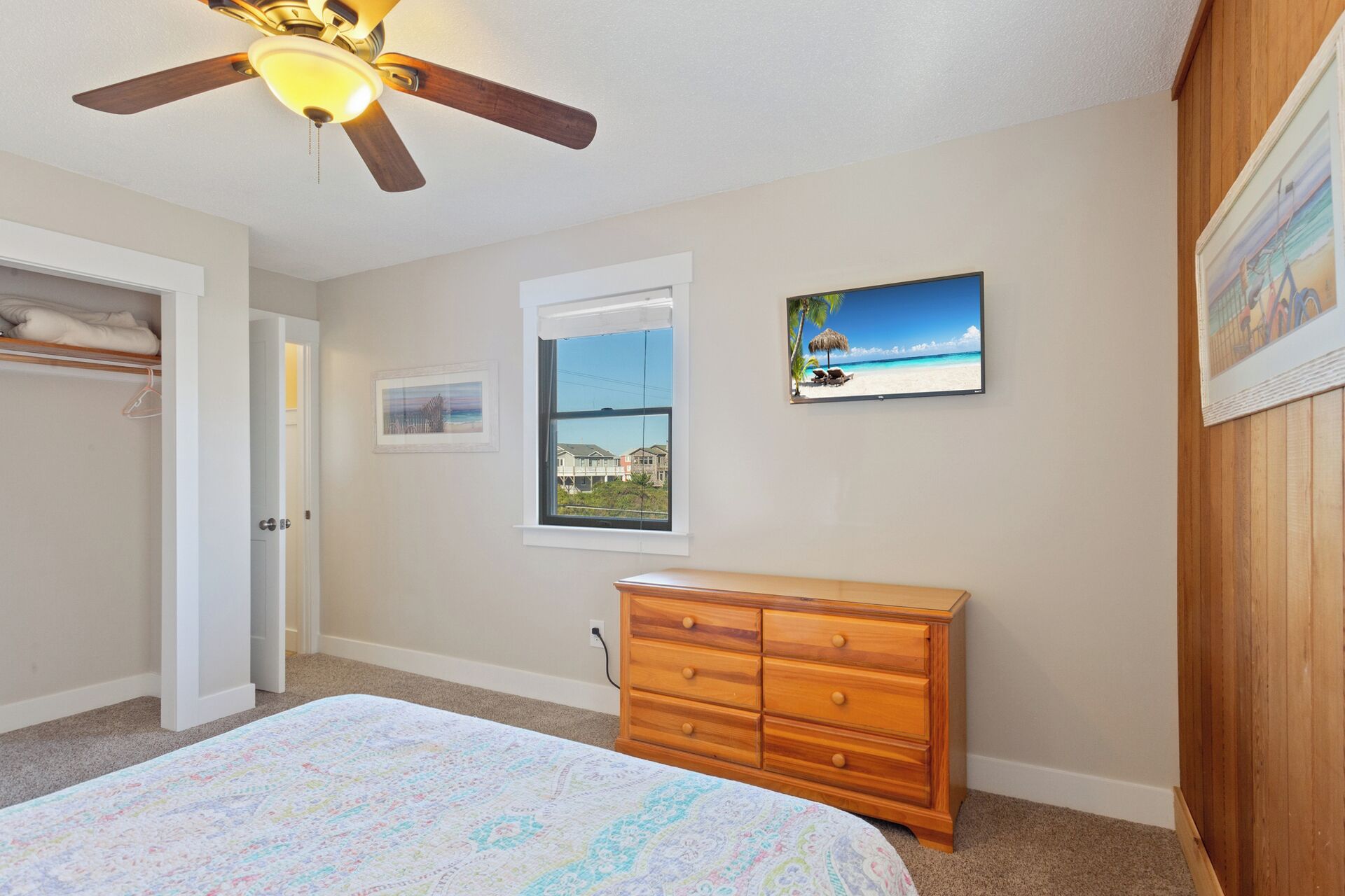 A bedroom with a bed, a dresser, a ceiling fan, a window with a view of a beach, and a framed picture on the wall.
