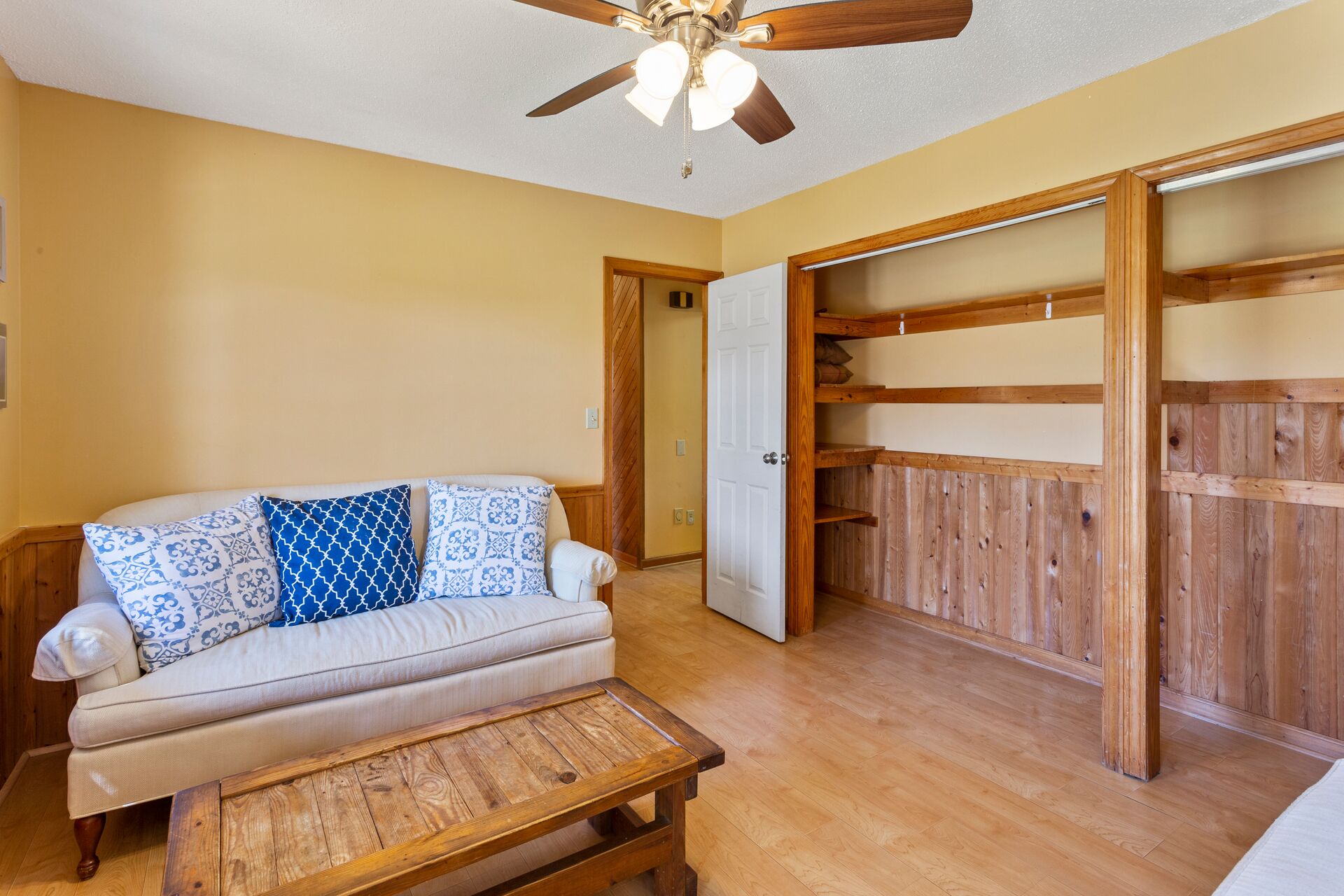 A room with a beige sofa, patterned cushions, a wooden coffee table, a ceiling fan, and a wooden closet with open doors.