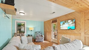 A cozy living room with a wooden ceiling, a teal wall, a large window, a television, and a patterned armchair. - Beach, Sunrise, Views, community pool! (Nags Head)