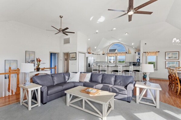 A spacious living area with a grey leather sectional sofa, a wooden coffee table, and a dining area with white chairs and a wooden table.